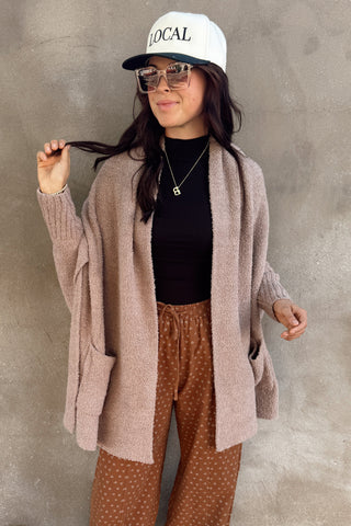 Person wearing a pink cardigan, black top, brown patterned pants, and white cap against a concrete wall.