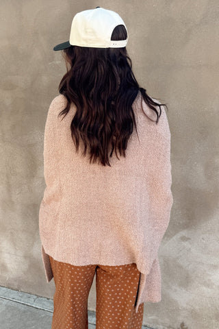 Person wearing a pink sweater and brown pants against a beige wall.