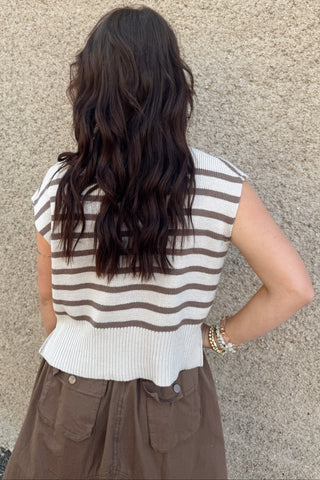 Person wearing a striped sleeveless top and brown pants against a beige wall.