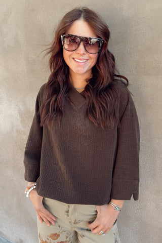 Woman wearing a brown sweater and sunglasses against a plain wall