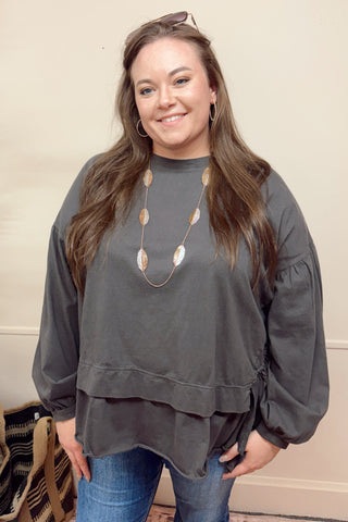 Woman wearing a gray layered top with long sleeves and a necklace, standing indoors.