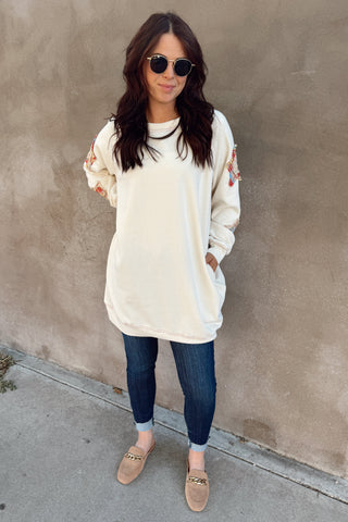Woman wearing a cream oversized sweatshirt, dark jeans, and sunglasses against a beige wall.