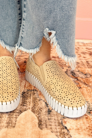Beige perforated sneakers with white soles worn with ripped jeans on a patterned floor.