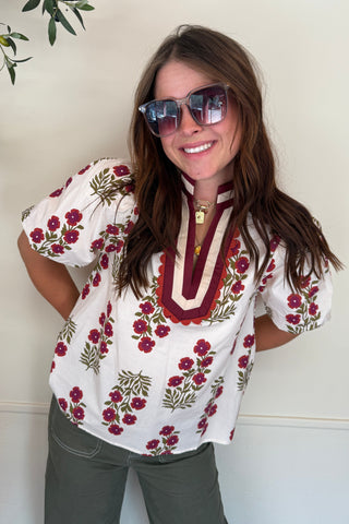 Woman wearing a floral blouse and sunglasses indoors with plants in the corner.