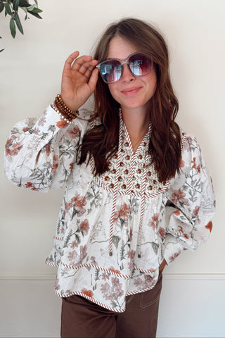 Woman wearing a floral blouse and sunglasses against a white wall.