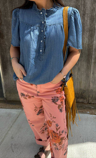 A person wearing a blue short-sleeve button-front blouse and pink floral pants, carrying a yellow fringe shoulder bag, standing outdoors against a wooden wall.