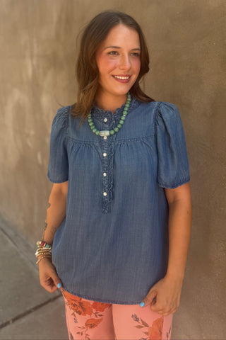 A woman wearing a short-sleeve blue chambray top with puff sleeves and a button-down front, paired with pink floral pants and a green beaded necklace, standing against a textured brown wall.