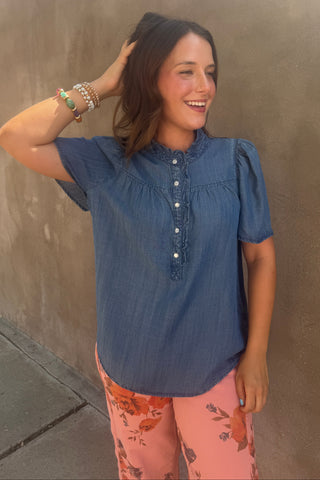 A woman wearing a blue chambray short-sleeve blouse with a buttoned mock neck and puff sleeves, paired with pink floral pants, posing against a textured brown stucco wall.