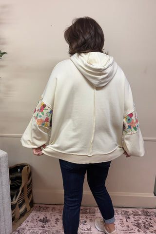 Person wearing a cream-colored jacket with floral embroidery in a room with a plant and sofa.