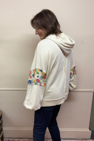 Person wearing a white hoodie with colorful designs in a room with a plant and rug.