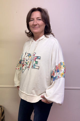 Person wearing a colorful hoodie with text in a room with a plant and furniture.