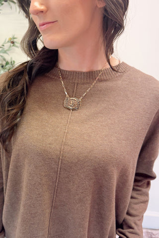 Woman wearing a brown sweater and gold necklace with a visible 'E' pendant.