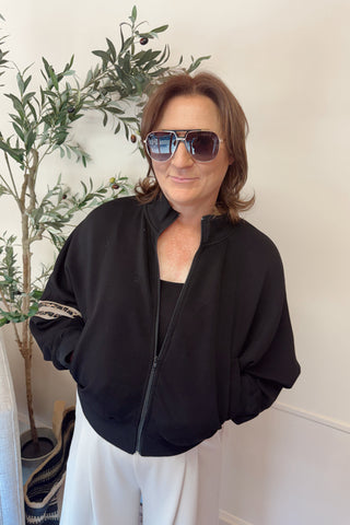 Person wearing a black bomber jacket and sunglasses indoors with a plant in the background