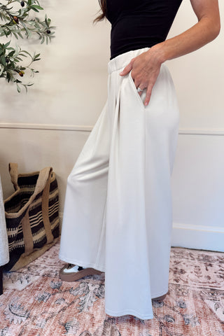 Person wearing white wide-leg pants and a black top in a room with a plant and bag in the background.