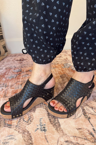 Black woven sandals worn with navy pants on a patterned rug