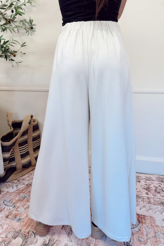 Person wearing a long white skirt in a room with a plant and rug.