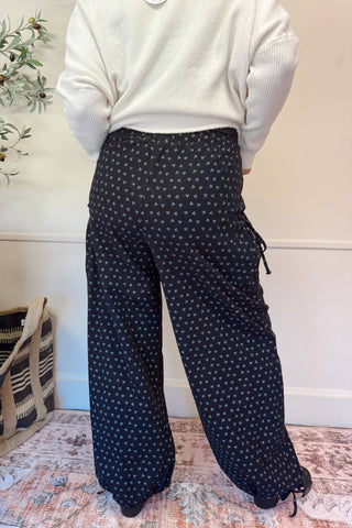 Person wearing black patterned pants and a white top in a room with a plant and furniture.