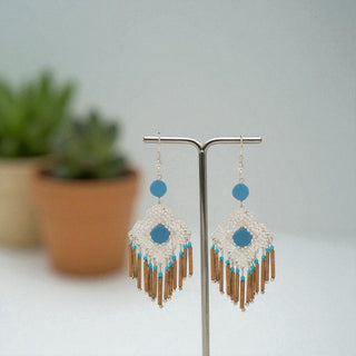 Pair of earrings with blue and gold design on a stand with a blurred plant background