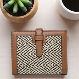 Small woven wallet with brown leather accents on a light surface with a plant and cup in the background